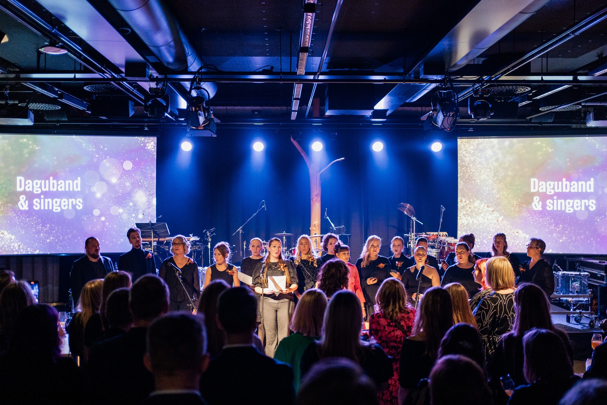 Dagmarin 50-vuotisjuhlissa laulanut dagmarilaisten oma kuoro lavalla, Daguband & singers teksti taustalla isoilla näytöillä